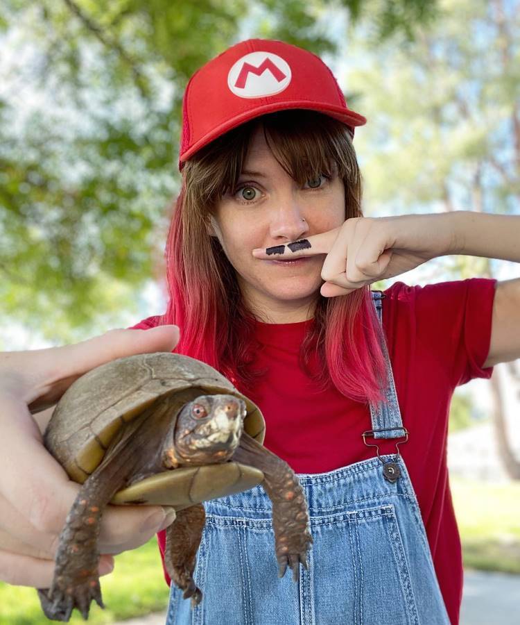 A imagem mostra uma mulher com uma divertida fantasia feminina para o Halloween, inspirada no Super Mario. Ela usa um boné vermelho com o símbolo do Mario, uma camiseta vermelha e um macacão jeans. Para completar o visual, ela tem um bigode falso desenhado no dedo, que segura de forma humorística sobre o lábio. Na mão, segura uma pequena tartaruga, remetendo aos icônicos inimigos do jogo. O ambiente ao ar livre, com árvores ao fundo, adiciona um toque natural à cena.