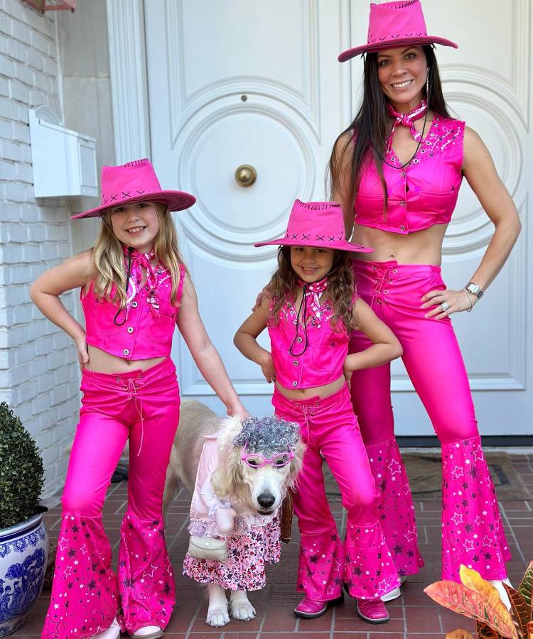 A imagem mostra três pessoas e um cachorro, todos vestidos com trajes de cowboy rosa brilhante. As duas crianças e a mulher usam chapéus, coletes e calças com detalhes em estrelas. O cachorro veste uma roupa rosa com óculos e uma peruca encaracolada, completando o visual divertido e coordenado. Eles estão posando em frente a uma porta branca, transmitindo uma sensação de alegria e descontração.