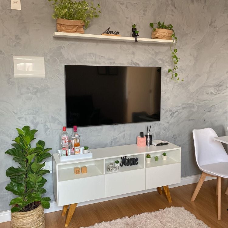 Foto de sala com parede de cimento queimado, televisão, rack branco, plantas na prateleira, vaso de palha com Ficus lyrata, piso de madeira e cadeira branca ao fundo do lado direito 