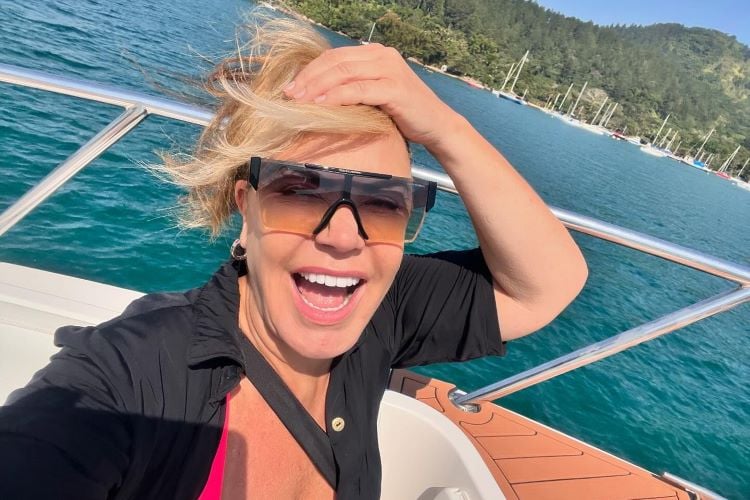 A imagem mostra Flor Fernandez sorridente em um barco. Ela está usando óculos escuros e segurando o cabelo com uma das mãos. O fundo apresenta um cenário de água azul e vegetação ao longe, com algumas embarcações ancoradas. O dia parece ensolarado.