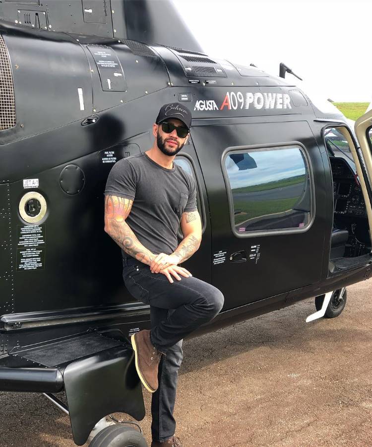 Gusttavo Lima com barba, tatuagens e óculos de sol está encostado em um helicóptero preto Agusta A109 Power. Ele usa camiseta preta, calça jeans escura, botas marrons e boné preto. O cenário é ao ar livre, com grama verde ao fundo. A imagem transmite uma sensação de estilo e aventura.