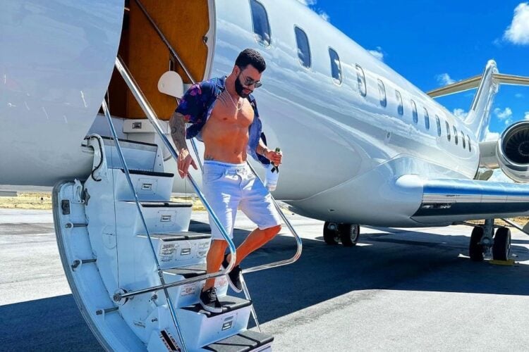 Gusttavo Lima desce as escadas de um jato particular, vestindo uma camisa aberta e shorts brancos. Ele segura uma garrafa na mão e usa óculos de sol. O céu está claro e azul, e a cena transmite uma sensação de luxo e lazer, com o jato estacionado em uma pista de aeroporto.