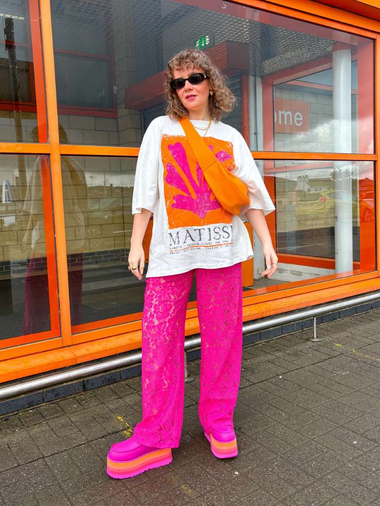 Mulher de pele clara usando camiseta branca, rosa e laranja, bolsa laranja, calça de renda rosa e sapato rosa 