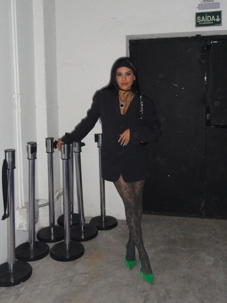 Mulher de pele morena usando blazer preto fechado, meia-calça de renda preta e sapato verde