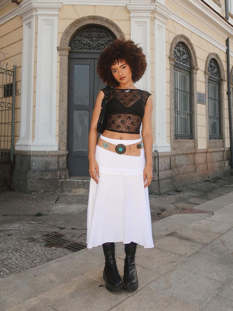 Mulher de pele negra usando cropped de renda preta, saia midi branca, botas e cinto bege 