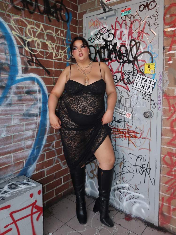 Mulher de pele clara usando vestido de renda preto transparente e botas pretas, com pichações ao fundo 