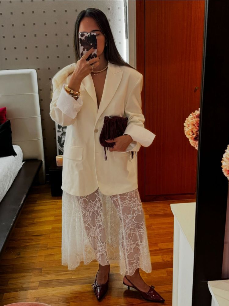 Mulher de pele clara tirando foto no espelho, usando blazer off white, saia branca de renda, bolsa pequena e sapato vinho 