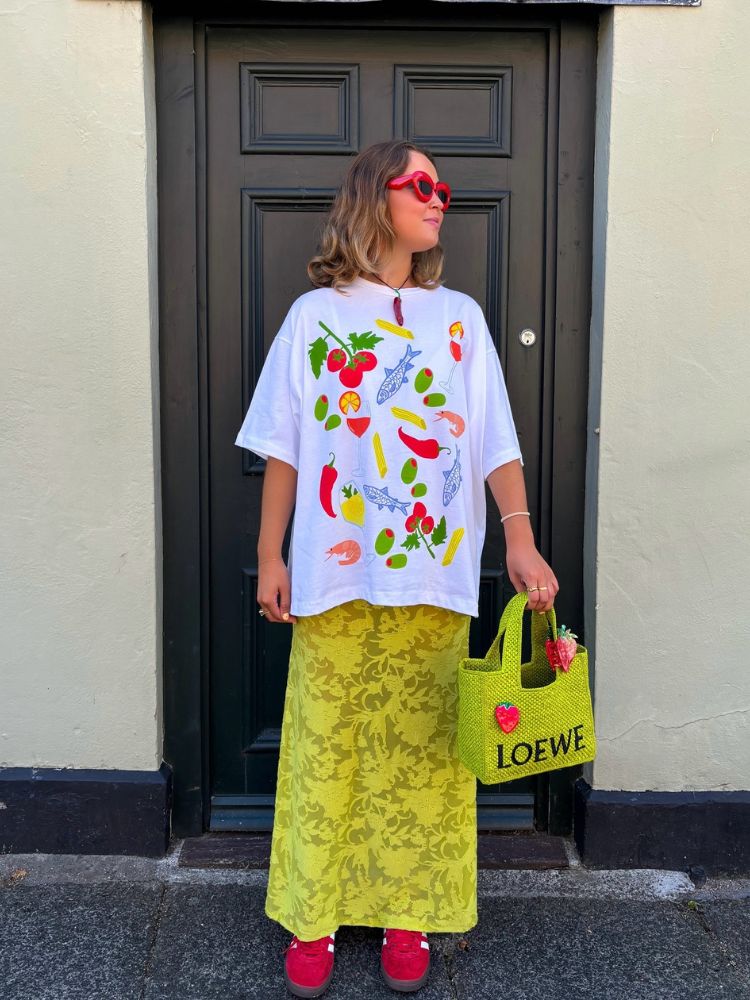 Mulher de pele clara usando camiseta branca com desenhos coloridos, saia amarela de renda, bolsa verde e tênis rosa 