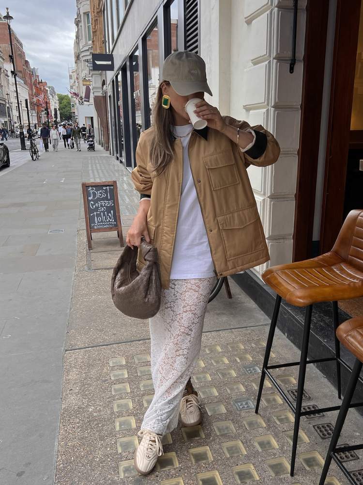 Mulher de pele clara em pé tomando café, usando bolsa marrom, saia bege, camiseta branca, jaqueta de couro bege e boné bege 