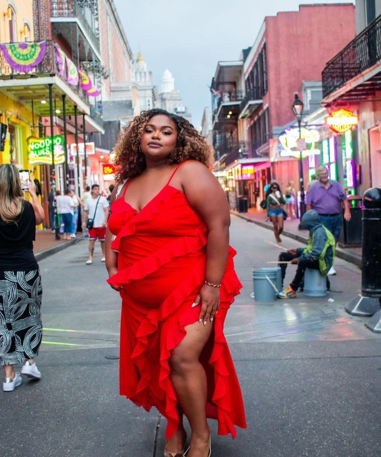 A imagem mostra uma mulher em uma rua movimentada, vestindo um deslumbrante vestido vermelho com babados e alças finas. O vermelho vibrante do vestido é uma cor da primavera 2024, destacando-se pela sua intensidade e elegância. Ao fundo, há prédios com luzes de neon e várias pessoas caminhando, criando um ambiente urbano e animado. A mulher tem cabelos cacheados e usa sandálias douradas, completando o visual sofisticado.