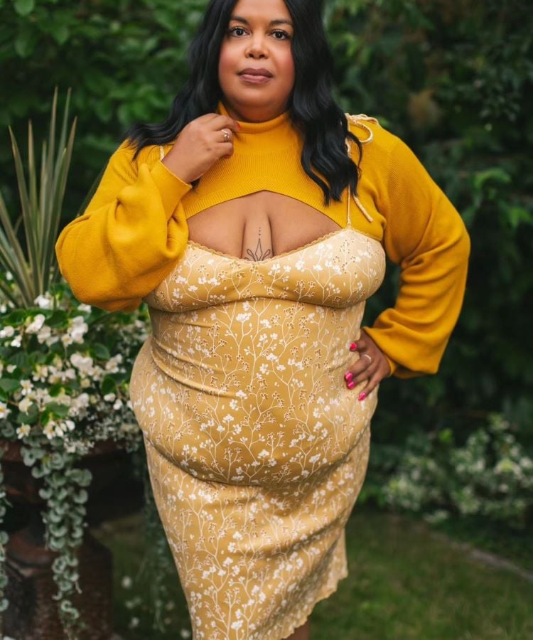 A imagem mostra uma mulher usando um vestido amarelo mostarda com estampa floral delicada, combinado com uma blusa de mangas longas na mesma tonalidade. O amarelo mostarda é uma cor da primavera 2024, destacando-se pela sua vivacidade e conexão com a natureza ao fundo.