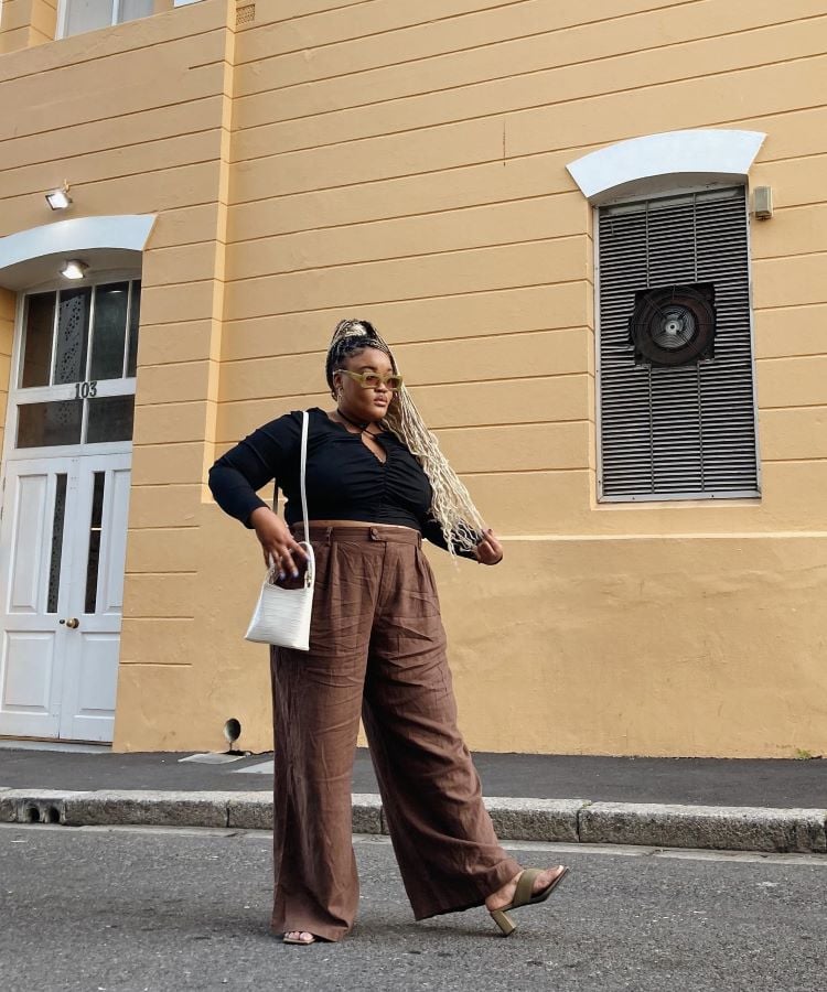 A imagem mostra uma mulher em pé na rua, em frente a um prédio de cor amarela. Ela está vestindo uma blusa preta de mangas compridas e calças largas marrons. A mulher usa sandálias de salto e carrega uma bolsa branca no ombro. Seu cabelo está estilizado em tranças longas, e ela usa óculos de sol. O ambiente urbano complementa o estilo moderno do visual.