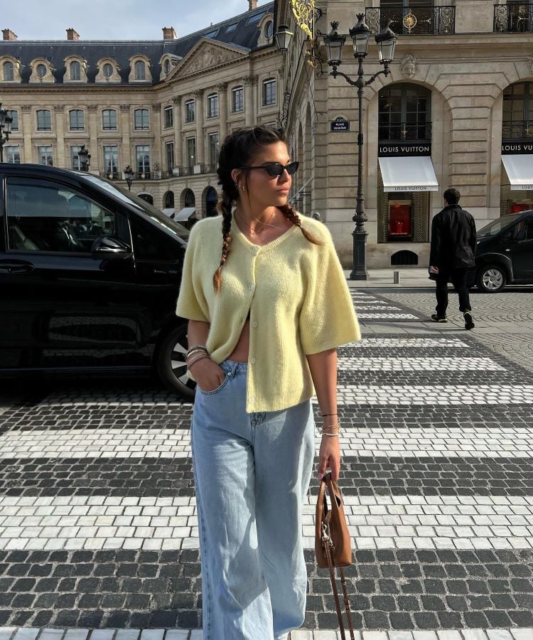 A imagem mostra uma mulher em uma rua elegante, vestindo um cardigã amarelo claro, que é uma cor da primavera 2024, trazendo um toque de suavidade e frescor. Ela combina o cardigã com calças jeans largas e segura uma bolsa marrom. A mulher usa óculos escuros e tem tranças, enquanto o cenário ao fundo inclui prédios clássicos e um carro preto estacionado.