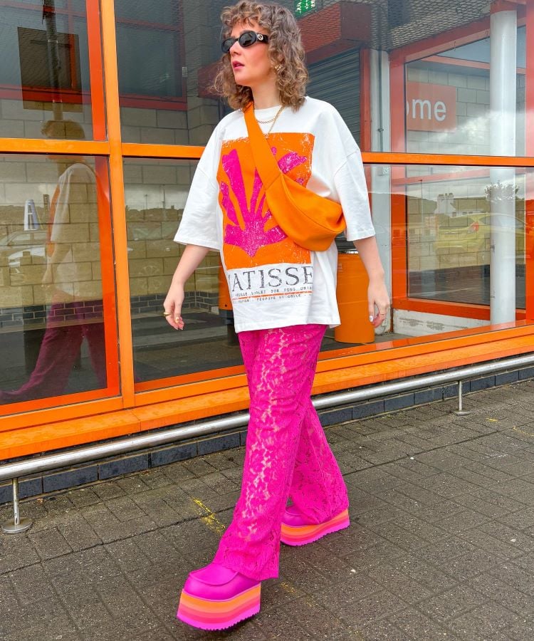 A imagem mostra uma mulher caminhando em frente a uma fachada de vidro e tijolos. Ela usa uma camiseta branca com uma estampa vibrante em rosa e laranja, combinada com calças rosa com textura e sapatos plataforma rosa com solado em camadas coloridas. A bolsa transversal laranja completa o look, destacando-se como uma cor da primavera 2024, trazendo um toque moderno e ousado ao visual. Ela também usa óculos de sol e tem cabelos cacheados.