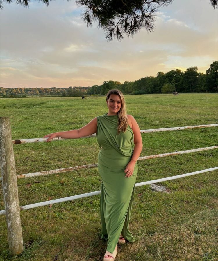 A imagem mostra uma mulher em um campo verde, vestindo um longo vestido verde oliva. Este tom de verde é uma cor da primavera 2024, trazendo uma sensação de frescor e conexão com a natureza. Ela está encostada em uma cerca de madeira, com árvores e um céu ao entardecer ao fundo, criando um cenário tranquilo e harmonioso.