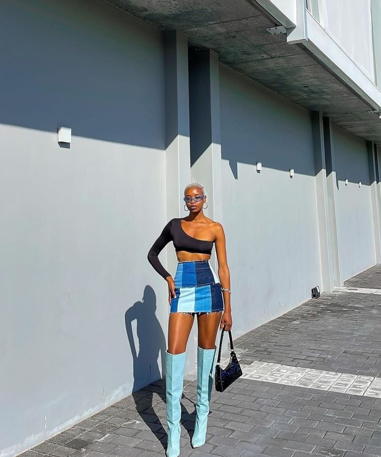 A imagem mostra uma mulher em pé em uma calçada, usando um top preto de uma manga e uma saia curta de patchwork jeans em tons de azul. Ela complementa o look com botas altas azuis e óculos de sol. A saia é uma inspiração para transformar calça jeans em saia, destacando estilo e criatividade.