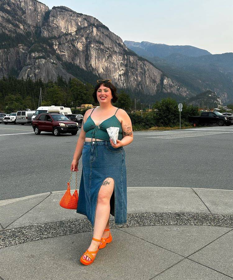 A imagem mostra uma mulher sorridente em frente a uma paisagem montanhosa. Ela usa um top verde e uma saia jeans longa com fenda, destacando uma tatuagem na perna. Ela segura uma bolsa laranja e um lanche. A saia é uma inspiração para transformar calça jeans em saia.