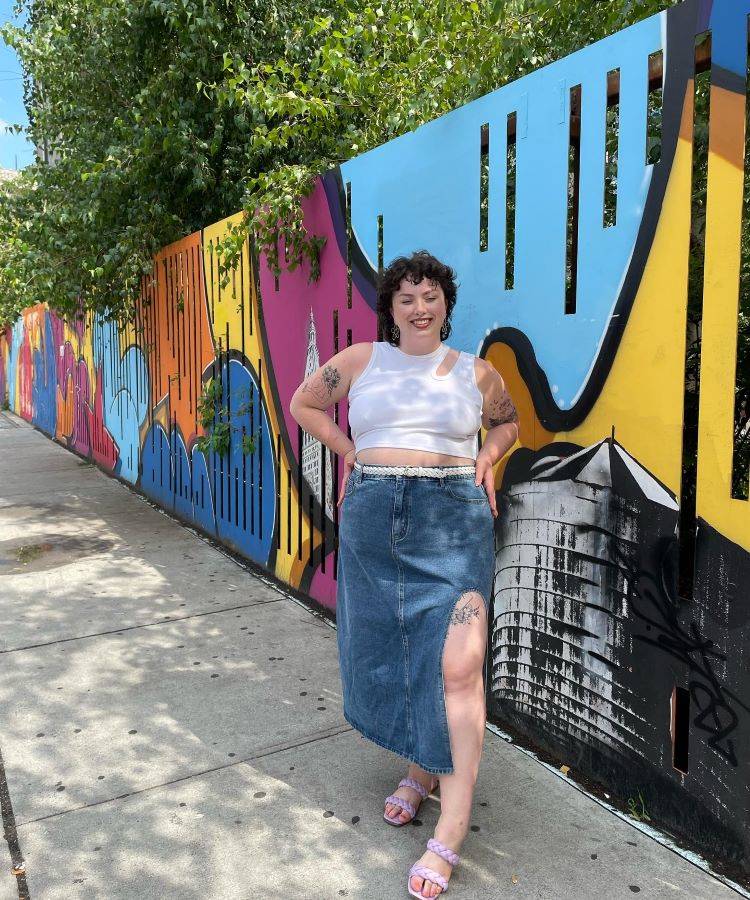 A imagem mostra uma mulher sorridente em pé na calçada, em frente a um mural colorido. Ela usa uma blusa branca sem mangas e uma saia jeans longa com fenda. Nos pés, sandálias lilás. O mural tem formas abstratas e cores vibrantes, como azul, amarelo e rosa.