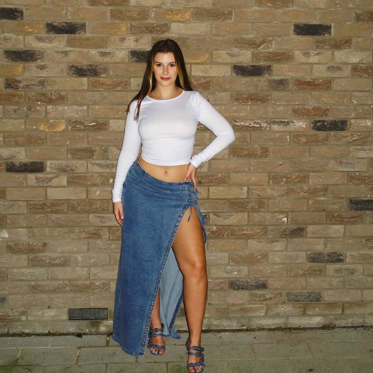 A imagem mostra uma mulher posando contra uma parede de tijolos, usando uma blusa branca de mangas longas e uma saia jeans longa com uma fenda alta na lateral. Ela está de sandálias azuis. O visual serve como inspiração para transformar calça jeans em saia, criando um estilo moderno e ousado.