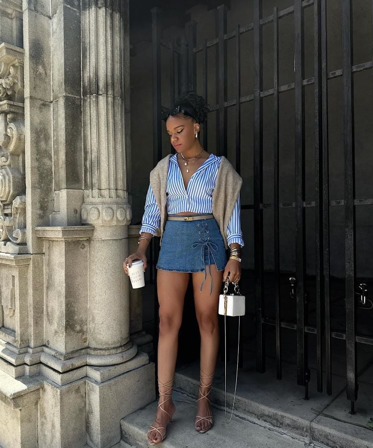 A imagem mostra uma mulher em frente a uma estrutura de pedra, vestindo uma camisa listrada azul e branca, com um suéter nos ombros. Ela usa uma saia jeans curta com amarração frontal e sandálias de tiras. Segura um copo e uma bolsa pequena. É uma inspiração para transformar calça jeans em saia.