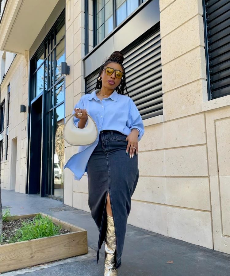 A foto mostra uma mulher em pé na calçada, usando uma camisa azul clara e uma saia jeans preta longa com uma fenda frontal. Ela segura uma bolsa branca e usa botas douradas. O visual é uma inspiração para transformar calça jeans em saia, criando um estilo elegante e moderno.