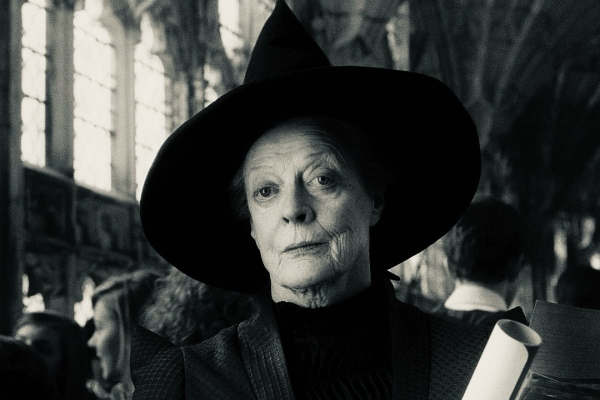 A imagem mostra a atriz Maggie Smith usando um chapéu pontudo, típico de uma bruxa, em um ambiente que parece ser uma construção antiga com grandes janelas ao fundo. A expressão facial é séria e a imagem está em preto e branco, o que dá um ar clássico e misterioso à cena.