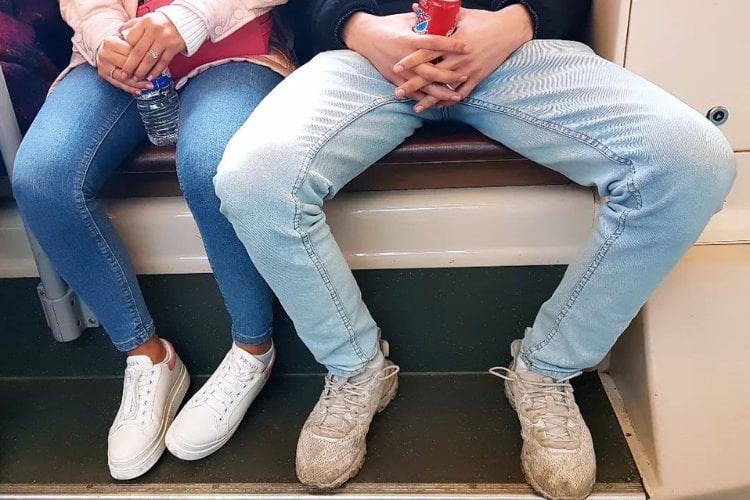 Homem de calça jeans com pernas abertas em assento, deixando mulher também de calça jeans sem espaço