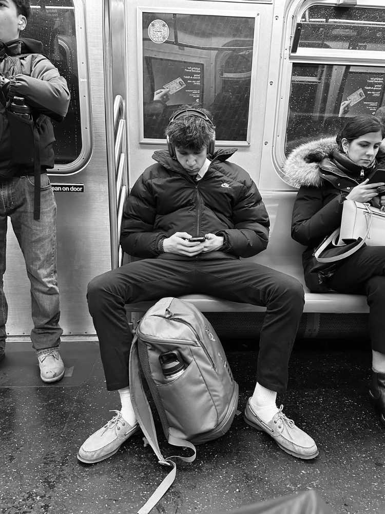 Foto em preto e branco de homem usando jaqueta, calça e com mochila no chão ocupando mais de assento. A mulher ao lado dele é obrigada a sentar tomando distância