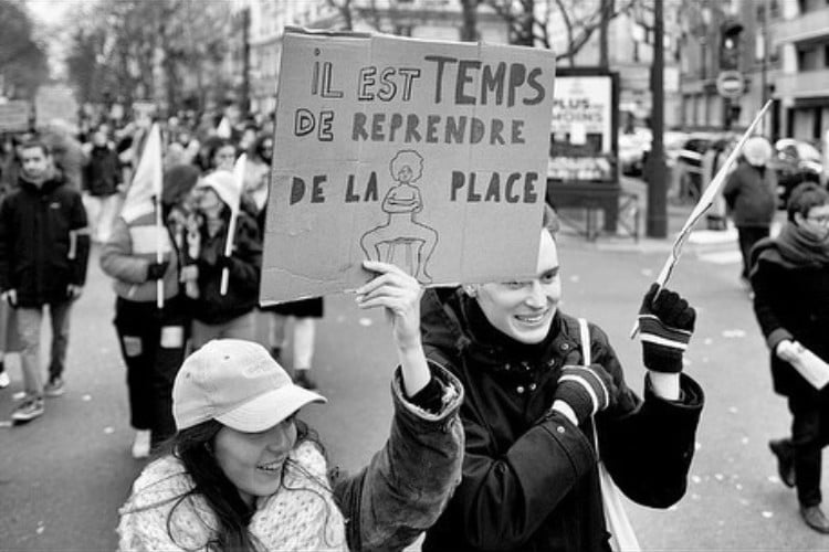 Mulheres protestando contra manspreading, com cartaz de papelão escrito "Il est temps de reprende de la place" e desenho de homem sentado com as pernas abertas 