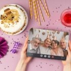 Foto de pessoa de pele clara segurando tablet com imagem de 4 idosos com chapéu dourado de festa. As mãos estão sobre mesa lilás que possui itens de festa, como bolo, vela, bexiga e confete