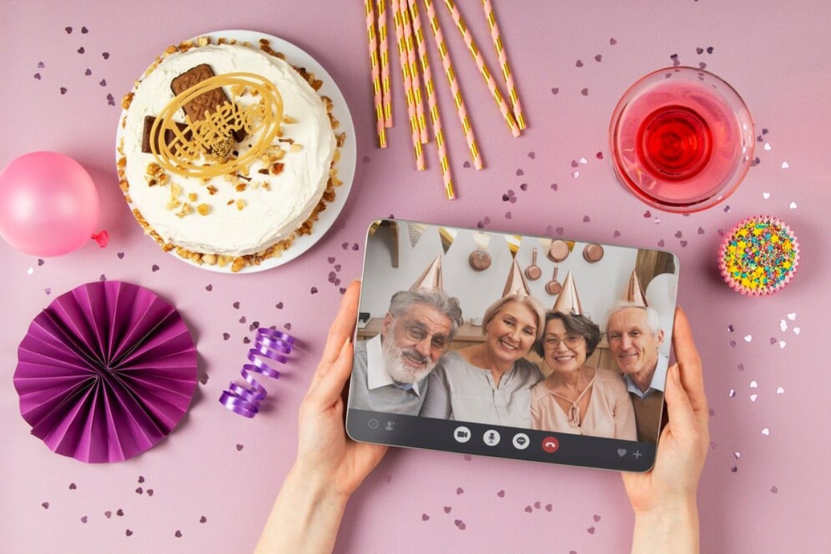 Foto de pessoa de pele clara segurando tablet com imagem de 4 idosos com chapéu dourado de festa. As mãos estão sobre mesa lilás que possui itens de festa, como bolo, vela, bexiga e confete