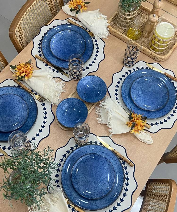 A imagem mostra uma mesa de jantar elegantemente arrumada. Há pratos azuis com bordas decorativas em sousplats brancos com detalhes em azul. Talheres dourados estão dispostos ao lado de guardanapos brancos com franjas, adornados com flores amarelas. Copos de vidro transparente estão ao lado dos pratos. No centro da mesa, há uma bandeja de palha com garrafas de vidro e um vaso com uma planta verde, criando um ambiente acolhedor e sofisticado. As cadeiras têm encostos de palha trançada.