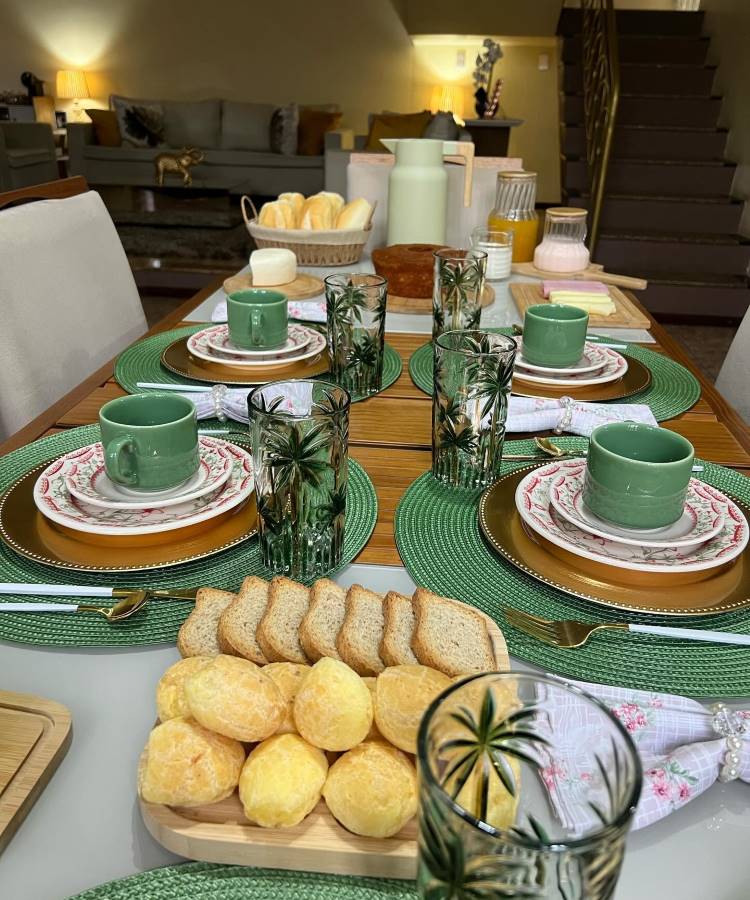 A imagem apresenta uma mesa posta para café da manhã, com jogos americanos verdes e pratos decorados com flores. As xícaras são verdes e estão acompanhadas por copos com desenhos de palmeiras. Talheres dourados e guardanapos brancos com detalhes florais completam a arrumação. No centro da mesa, há pães de queijo e torradas em uma tábua de madeira. Ao fundo, uma cesta com pães e jarras de suco e café adicionam ao ambiente acolhedor e convidativo.