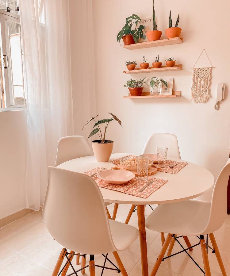 A imagem mostra uma sala de jantar iluminada por luz natural, com uma mesa redonda branca cercada por quatro cadeiras de design moderno, com assentos brancos e pernas de madeira. Sobre a mesa, há um vaso com uma planta e jogos americanos com pratos e copos transparentes. Na parede ao fundo, prateleiras de madeira exibem vasos de plantas em tons de terracota. Uma peça de macramê pendurada adiciona um toque decorativo, criando um ambiente acolhedor e minimalista.