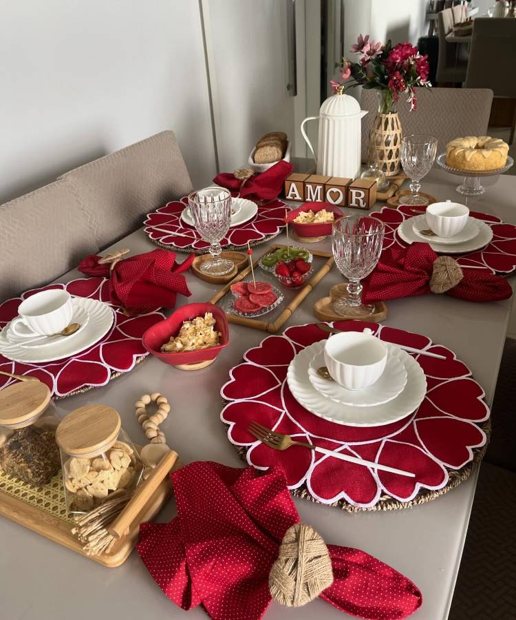 A imagem exibe uma mesa posta para café da manhã, destacando sousplats vermelhos em formato de coração. Pratos brancos e xícaras estão acompanhados de guardanapos vermelhos com porta-guardanapos de palha. Taças de vidro elegante completam a arrumação. No centro, há uma garrafa térmica branca e blocos de madeira formando a palavra "AMOR". Bandejas de madeira contêm biscoitos e pães, enquanto um prato elevado exibe um bolo. Um vaso com flores vermelhas adiciona charme ao ambiente, criando um cenário romântico e acolhedor.