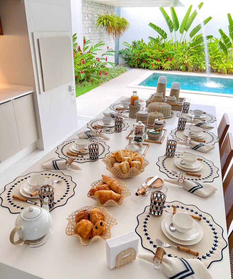 A imagem mostra uma mesa posta para café da manhã em um ambiente ao ar livre, próximo a uma piscina. Há sousplats com bordas decorativas azuis, pratos brancos e xícaras combinando. Talheres com cabos de bambu e guardanapos de tecido acrescentam elegância. No centro, cestas de pães e croissants estão dispostas ao lado de uma bandeja com suco e potes de geleia. Copos com detalhes geométricos completam a arrumação, criando um ambiente convidativo e sofisticado. Ao fundo, um jardim tropical adiciona frescor à cena.