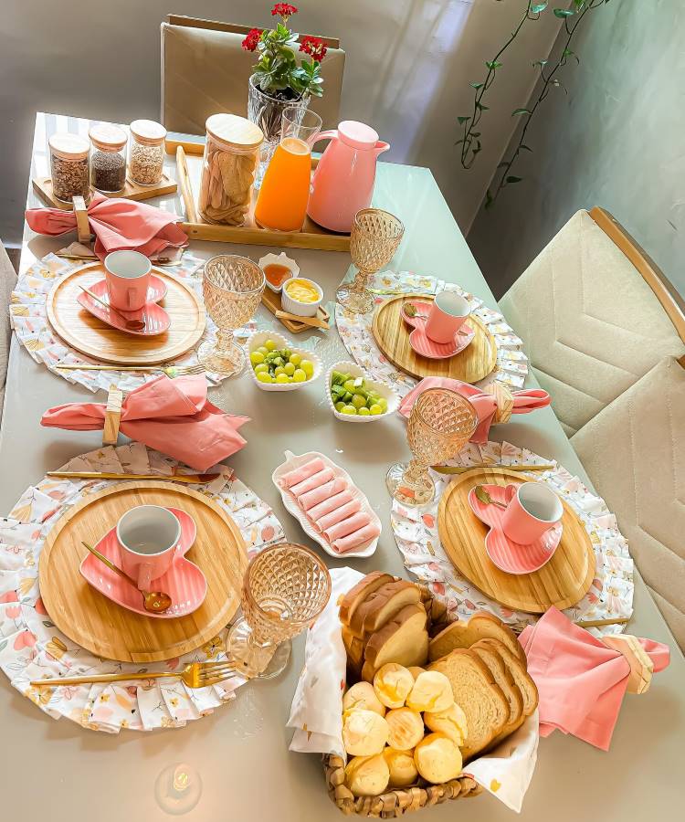 A imagem mostra uma mesa de café da manhã elegantemente posta. Há pratos de madeira com xícaras e pires rosa, cercados por guardanapos cor de rosa. Taças de vidro complementam o arranjo. No centro, há tigelas com uvas verdes e amarelas, além de uma bandeja com potes de cereais e uma jarra de suco laranja. Um cesto de pães variados e flores vermelhas em um vaso completam a decoração, criando um ambiente acolhedor e convidativo.