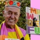 A imagem mostra um homem sorridente usando um chapéu colorido com bordados de flores. Ele veste uma camisa amarela com detalhes em rosa e laranja. Ao lado, há um livro intitulado "Mr. Wonderful" por Dininha Morgado. A capa do livro é vibrante, com letras grandes e coloridas, e uma foto em preto e branco do homem destacada. O fundo da imagem é dividido entre um cenário natural e uma cor rosa sólida.