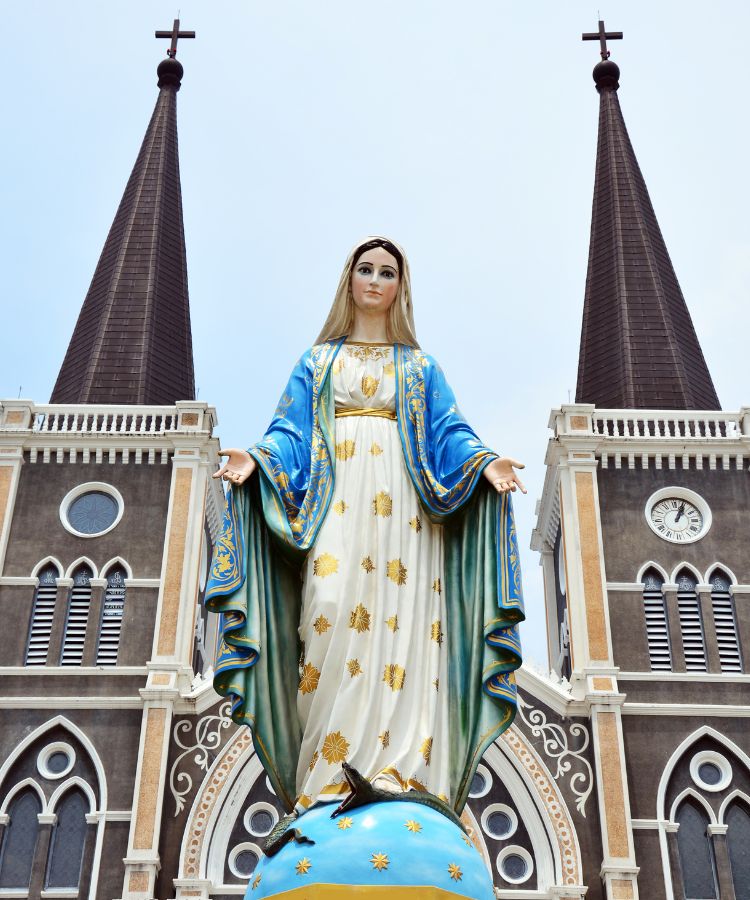 A imagem mostra uma estátua de Nossa Senhora em frente a uma igreja. A figura está de pé sobre um globo azul com estrelas douradas, vestindo um manto azul e branco decorado com estrelas douradas. Seus braços estão abertos. Ao fundo, há duas torres gêmeas da igreja, cada uma com uma cruz no topo.