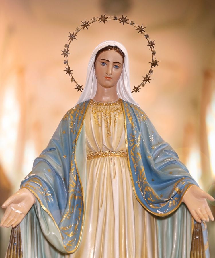 A imagem mostra uma estátua de Nossa Senhora com um olhar sereno. Ela veste um manto azul com detalhes dourados e uma túnica branca. Seu véu é branco, e um halo de estrelas circunda sua cabeça. As mãos estão estendidas para frente. O fundo é suave e desfocado, destacando a figura central.