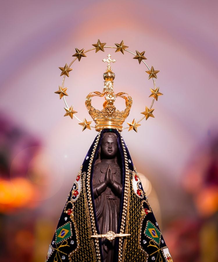 A imagem mostra a estátua de Nossa Senhora Aparecida, padroeira do Brasil. A figura é escura, com mãos postas em oração, vestida com um manto azul escuro adornado com bordados dourados e bandeiras do Brasil. Ela usa uma coroa dourada, e um halo de estrelas douradas circunda a coroa. O fundo é desfocado, destacando a estátua.