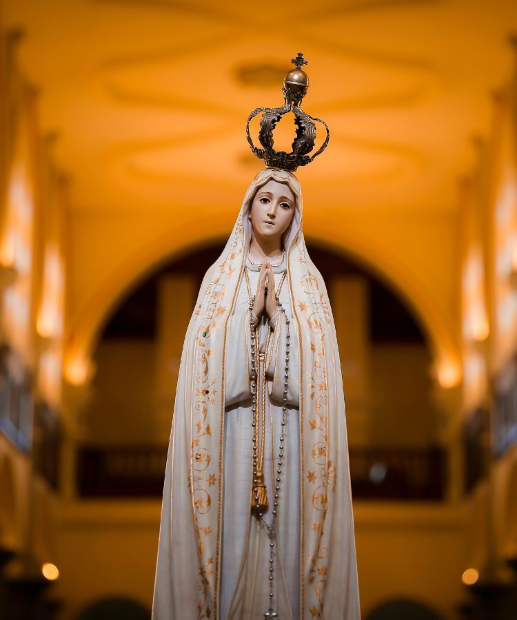 A imagem mostra uma estátua de Nossa Senhora de Fátima. Ela está vestida com um manto branco decorado com bordados dourados e segura um rosário. Suas mãos estão em oração, e ela usa uma coroa dourada. O fundo é uma igreja com iluminação suave, destacando a figura central. A expressão da estátua é serena e contemplativa.