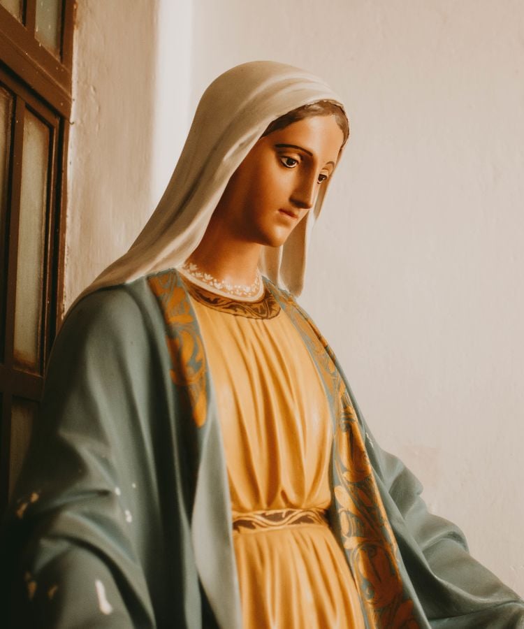 A imagem mostra uma estátua de uma mulher com expressão serena, vestida com um manto azul esverdeado e uma túnica amarela. Ela usa um véu branco sobre a cabeça. A estátua está posicionada em um ambiente interno, próximo a uma janela de madeira, com luz suave iluminando o rosto e as vestes detalhadas.