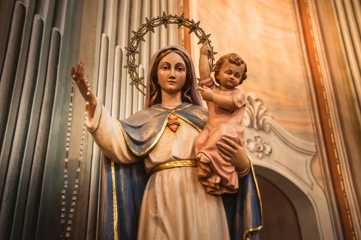 A imagem mostra uma estátua de uma mulher segurando uma criança no colo. A mulher veste um manto azul e branco e tem um halo de estrelas ao redor da cabeça. Ela segura um rosário em uma das mãos. A criança está vestida de rosa e parece ser um menino. O fundo é detalhado e ornamentado.