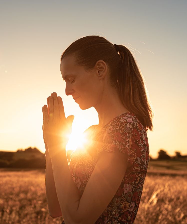 A imagem mostra uma mulher em perfil, de olhos fechados, em posição de oração com as mãos unidas. Ela está em um campo ao pôr do sol, vestindo uma blusa estampada. A luz do sol brilha intensamente atrás dela, criando um efeito de halo ao redor de sua silhueta e destacando a serenidade do momento.