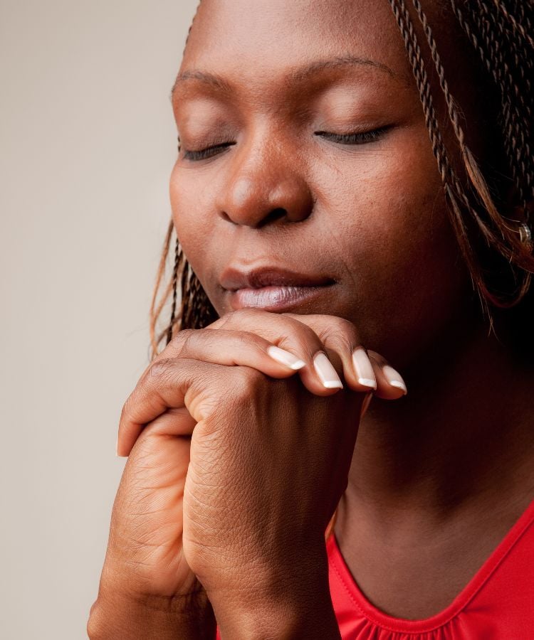 A imagem mostra uma mulher de olhos fechados em um momento de reflexão ou oração. Ela tem tranças finas e está vestindo uma blusa vermelha. Suas mãos estão unidas em frente ao rosto, apoiando o queixo. A expressão é serena e tranquila, com um fundo neutro que destaca sua presença.