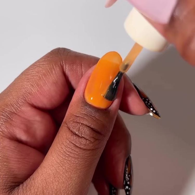 Aplicação de esmalte laranja na unha, criando a base para o design de borboleta.