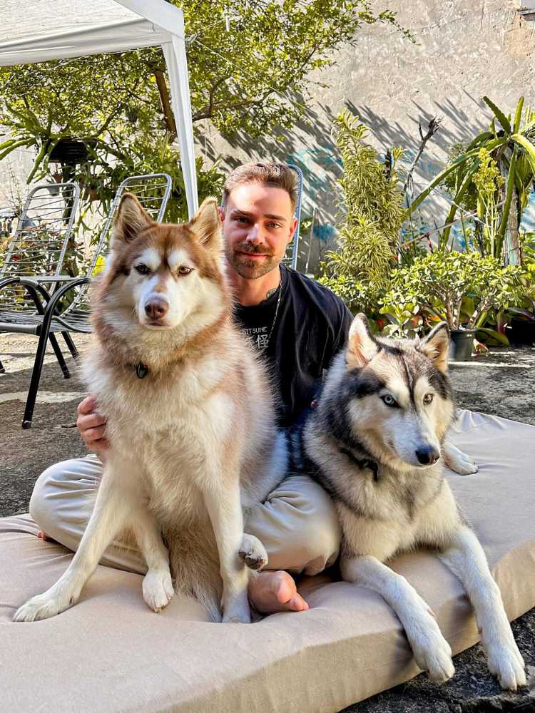 Zanq sentado usando blusa preta e calça cinza, abraçado com dois Huskies 