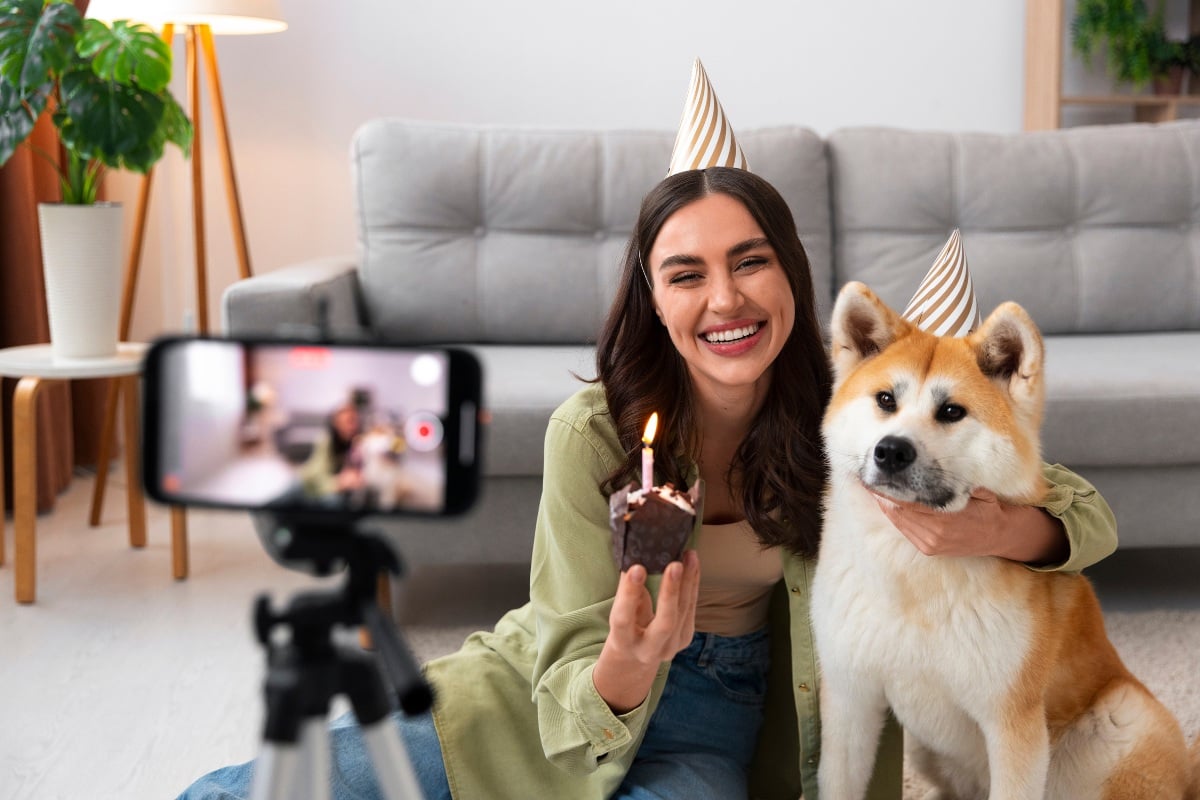 Foto de mulher de pele clara sentada em frente à sofá cinza abraçada com cachorro branco e alaranjado, ambos suando chapéu de aniversário e em frente à celular gravando em tripé