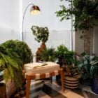 A imagem mostra um ambiente interno decorado com várias plantas em vasos, criando uma atmosfera aconchegante e natural. Há uma variedade de plantas, incluindo samambaias e outras espécies de folhagem, dispostas em vasos de diferentes tamanhos e padrões. Um banco acolchoado, de madeira clara com tiras de tecido, está posicionado no centro da cena, com um livro aberto e um par de óculos sobre ele, sugerindo um espaço de leitura ou relaxamento. Ao fundo, há uma luminária de chão com um design moderno, com uma haste curva e uma cúpula metálica dourada que ilumina suavemente o ambiente. As paredes são claras e há uma grande janela com cortinas translúcidas, permitindo a entrada de luz natural, o que contribui para o crescimento das plantas e reforça a sensação de tranquilidade do espaço. O piso tem um padrão geométrico em tons de preto, branco e cinza, complementando a decoração moderna e natural do ambiente.